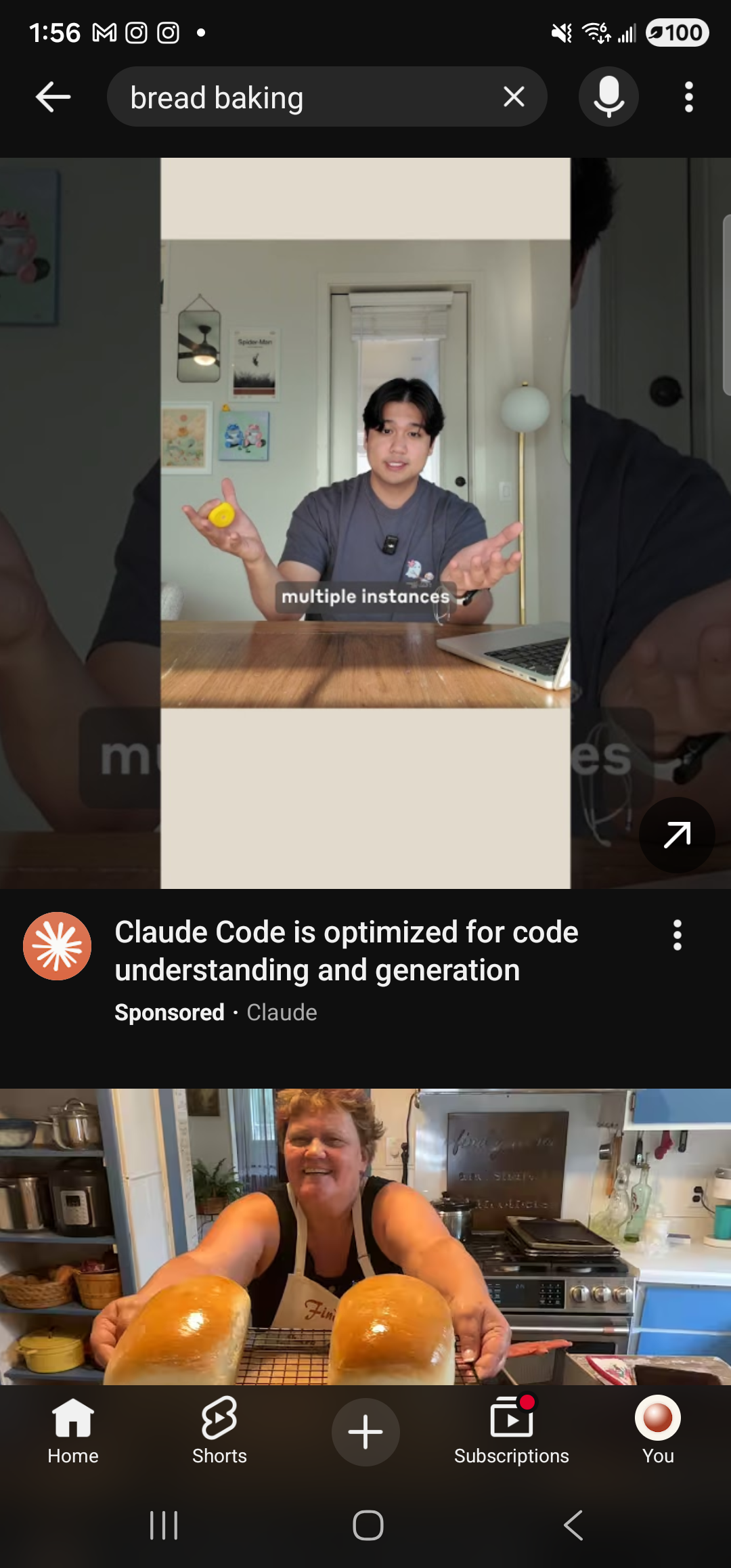 The YouTube Android app foregrounded on the same Samsung Galaxy S23 Ultra in dark mode. At the top, a back arrow, a dark search field showing the query ‘bread baking’ with an ✕ clear button, a microphone search icon, and a 3-dot overflow menu. Below the search bar is a tall portrait video tile — a person in a gray t-shirt seated at a kitchen table, holding two yellow round loaves on either side, with the text overlay ‘multiple instances’. Beneath the tile a clay-orange channel avatar, the title ‘Claude Code is optimized for code understanding and generation’, a ‘Sponsored · Claude’ label, and a 3-dot menu — a sponsored result. Below that, the top edge of a second video thumbnail showing a smiling person with two more golden loaves on a counter. The bottom navigation bar shows YouTube’s standard tabs Home, Shorts, a centered ‘+’ create button, Subscriptions, and You.