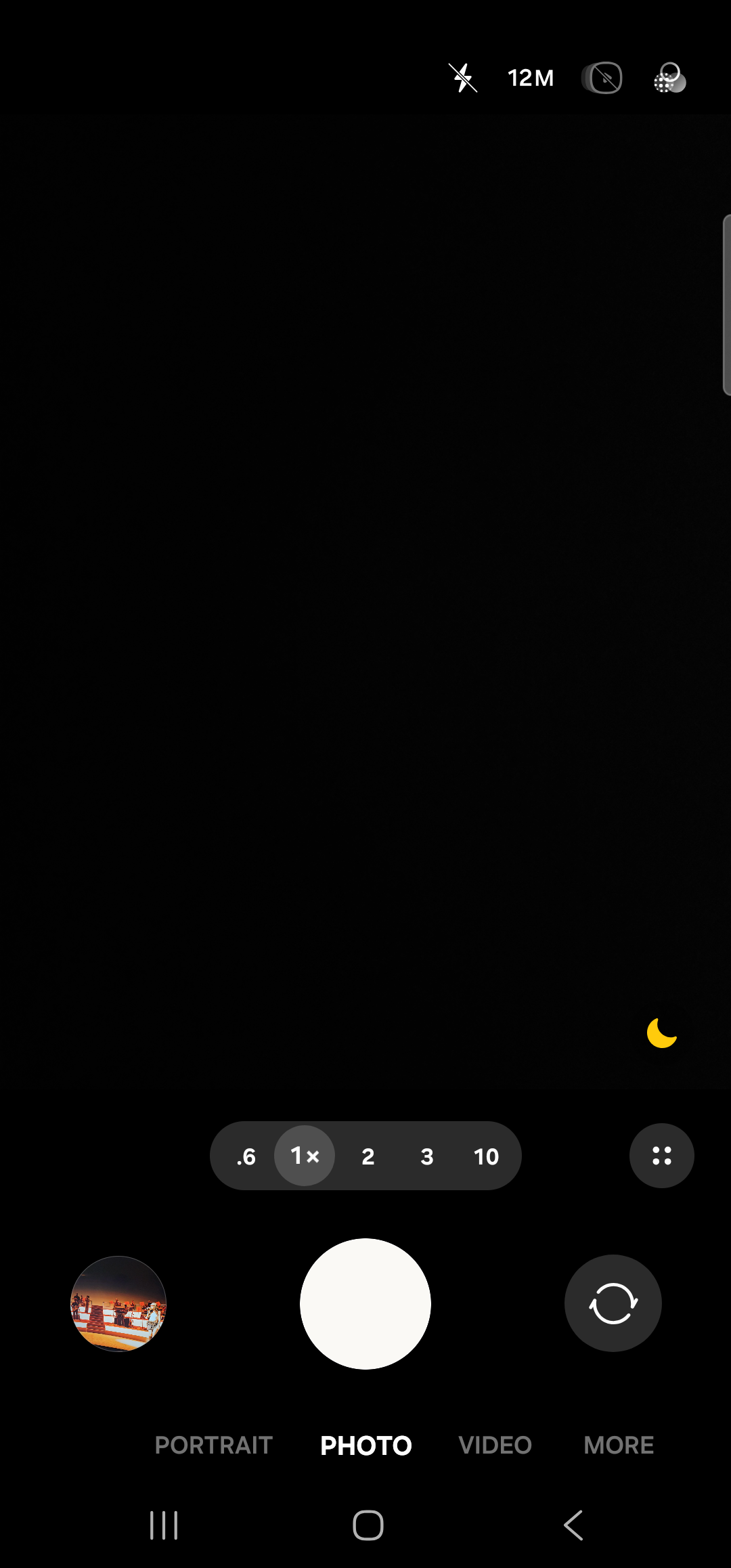 The Samsung default camera app foregrounded, in still-image mode on the rear lens. The viewfinder fills the screen, almost entirely black because the lens is covered. Across the top right are the flash icon, a ‘12M’ resolution badge, an aspect-ratio toggle, and a settings cog. Near the lower-right corner of the viewfinder is a small crescent-moon scene-mode icon. Below the viewfinder a horizontal row of zoom chips reads ‘.6  1×  2  3  10’, with 1× selected. Below that, the standard camera control row: a thumbnail of the most recent photo on the left, a large white shutter button in the center, and a camera-flip icon on the right. Beneath the controls a row of mode tabs reads ‘PORTRAIT  PHOTO  VIDEO  MORE’, with PHOTO highlighted. The Samsung 3-button nav sits at the very bottom.