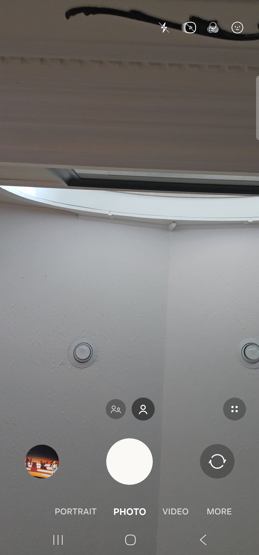 The Samsung default camera app foregrounded, in still-image mode with the front-facing lens active. The viewfinder fills the screen showing the room the phone is in — pale walls, a ceiling vent, and overhead lighting in soft focus, since the phone is lying on a desk facing up. Across the top right are the flash icon, an aspect-ratio toggle, a settings cog, and an effects icon. Two small selfie face/group icons sit just above the control row near the center. Below the viewfinder the standard camera control row: a thumbnail of the most recent photo on the left, a large white shutter button in the center, and a camera-flip icon on the right. Beneath the controls a row of mode tabs reads ‘PORTRAIT  PHOTO  VIDEO  MORE’, with PHOTO highlighted. The Samsung 3-button nav sits at the very bottom.