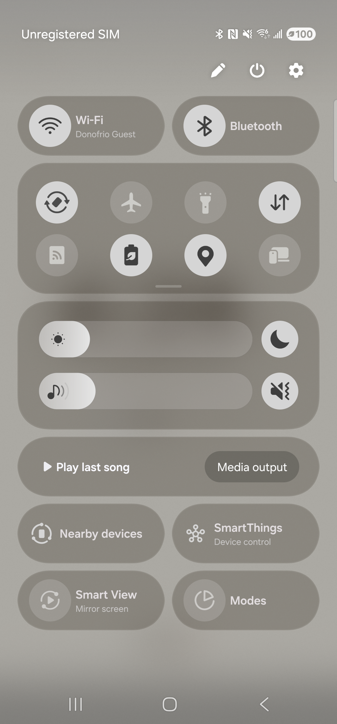 The Samsung One UI Quick Settings tray, fully expanded over a dimmed background. The top status row shows ‘Unregistered SIM’ on the left and Bluetooth / NFC / signal / Wi-Fi / battery icons on the right, with a pencil edit icon, a power button, and a settings cog at the very top-right of the tray. The tray is divided into rounded panels: a top panel with the Wi-Fi (Donofrio Guest) and Bluetooth tiles, a middle panel with a 2x4 grid of additional tiles (Auto rotate, Flight mode, Flashlight, Mobile data, Mobile Hotspot, Power saving, Location, Link to Windows), then a brightness slider and a media-volume slider, and finally cards for ‘Play last song / Media output’, ‘Nearby devices / SmartThings’, and ‘Smart View / Modes’. The pencil edit affordance at the top-right is the next interaction target.