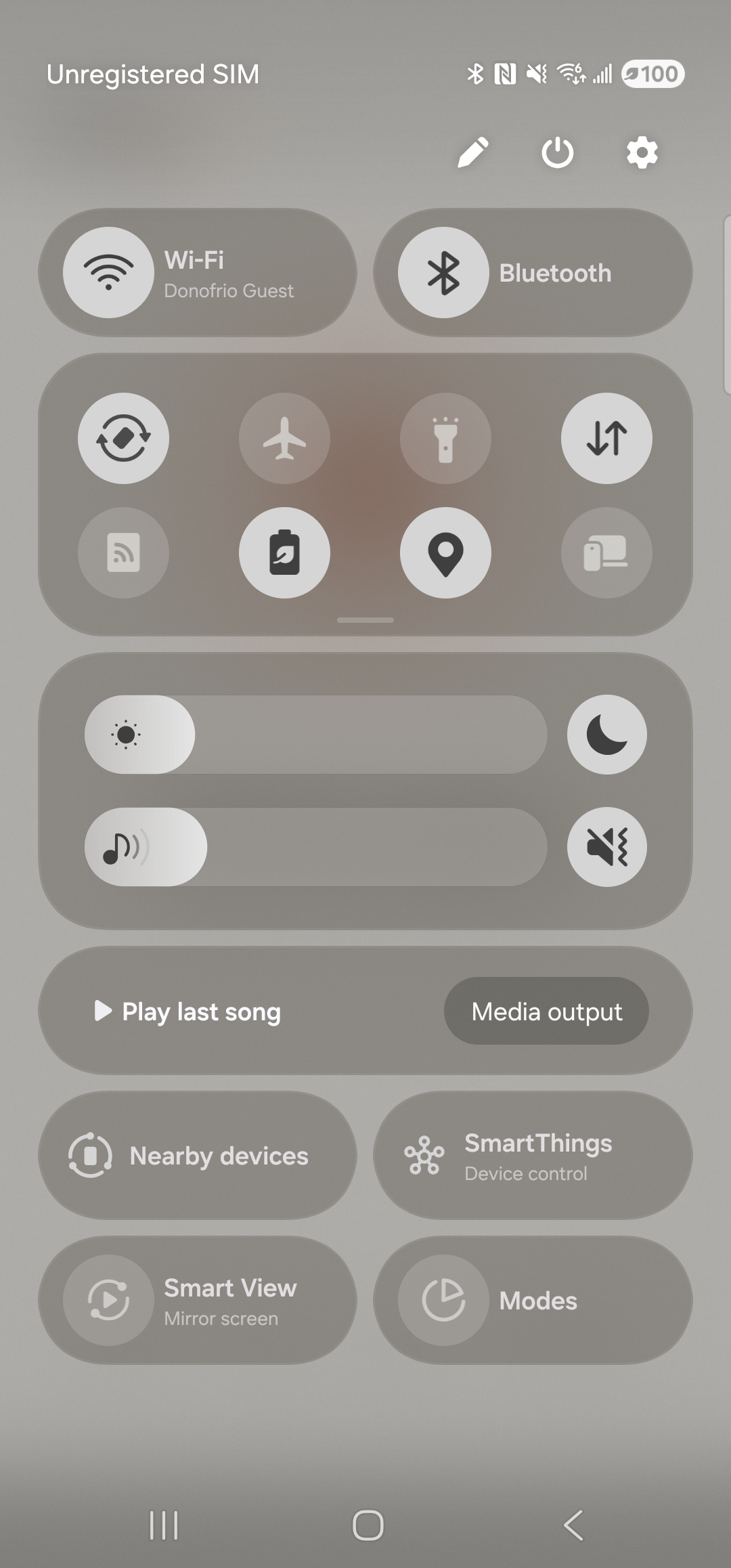 The Samsung One UI Quick Settings tray fully expanded on a Galaxy S23 Ultra, dimming the underlying app. The status row at the top shows ‘Unregistered SIM’ on the left and the usual signal/Wi-Fi/battery icons on the right. Top-right of the tray are three controls: a pencil (Edit buttons), a power button, and a settings cog. Below that, rounded panels group: a Wi-Fi (Donofrio Guest) and Bluetooth pair, a 2x4 grid of round tiles (Auto rotate, Flight mode, Flashlight, Mobile data, Mobile Hotspot, Power saving, Location, Link to Windows), brightness and media-volume sliders, and three media/device cards (‘Play last song / Media output’, ‘Nearby devices / SmartThings’, ‘Smart View / Modes’).